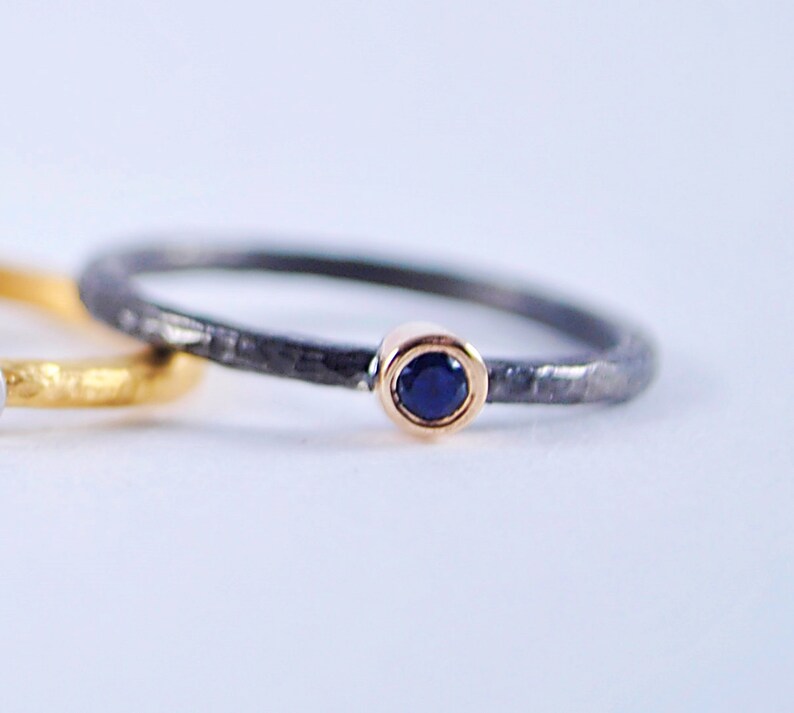 May include: A thin black ring with a hammered texture and a small blue gemstone set in a gold bezel.