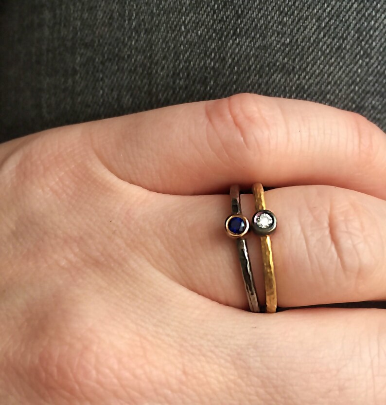May include: Two thin gold and black rings with small gemstones. The ring on the left has a blue gemstone and the ring on the right has a white gemstone.