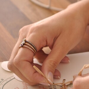 May include: Three gold rings with small gemstones. The rings are stacked on a finger. The gemstones are red, white, and yellow.