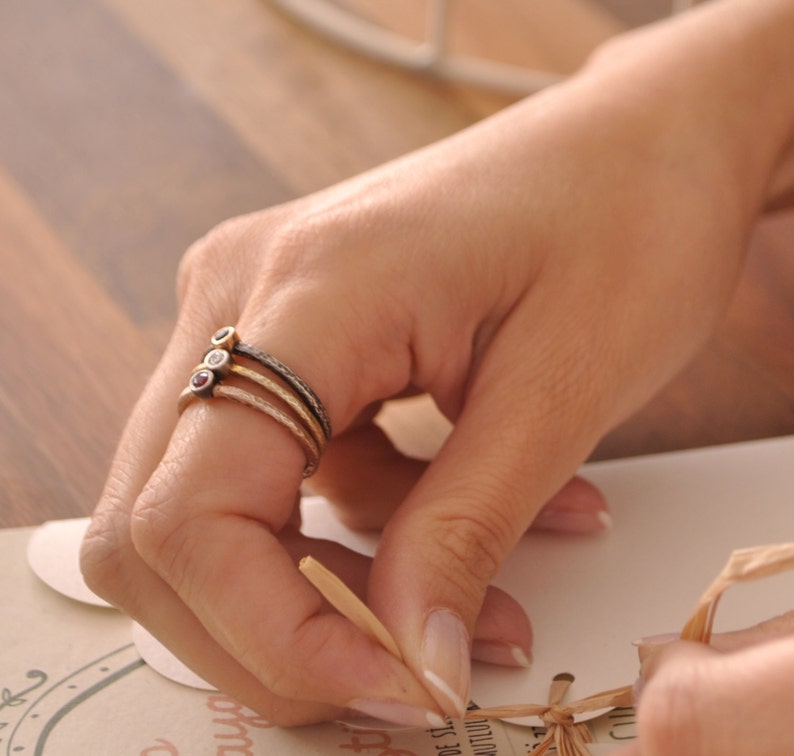 May include: Three gold rings with small gemstones. The rings are stacked on a finger. The gemstones are red, white, and yellow.