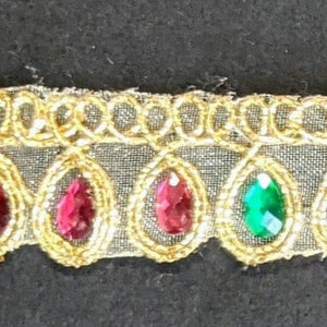 May include: Gold-colored trim with a repeating pattern of teardrop-shaped jewels in red, green, and blue. The jewels are set within gold-colored frames and attached to a sheer, light-colored fabric.