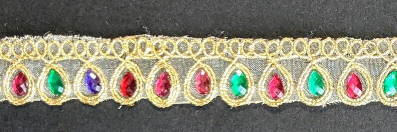 May include: Gold-colored trim with a repeating pattern of teardrop-shaped jewels in red, green, and blue. The jewels are set within gold-colored frames and attached to a sheer, light-colored fabric.