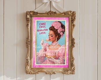 Può includere: Una stampa incorniciata con una cornice dorata ornata e un fiocco bianco in alto. La stampa raffigura una donna in abito rosa che mangia una torta, con il testo "CAKE FIRST, REVOLUTION LATER." Lo sfondo è azzurro.