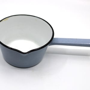 May include: A small, light blue enamel saucepan with a white interior and a black rim. The pan features a single long handle with a hole at the end for hanging and a small pouring spout on the side, designed for heating liquids or small portions.