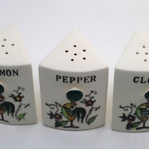 May include: Three white ceramic spice shakers with a rooster design. Each shaker is labelled with a spice name: Cinnamon, Pepper, and Cloves. The shakers have a triangular shape with holes on top for dispensing.
