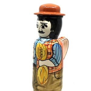 May include: A ceramic jug shaped like a person wearing a brown hat, blue and white striped shirt, red and white checked waistcoat, and brown trousers. The person is holding a yellow sheaf of wheat in their right hand. The jug has a brown handle.