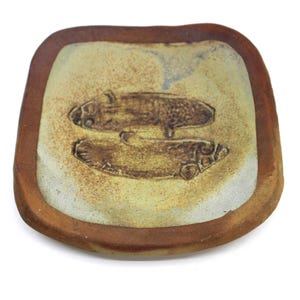 Peut inclure: Un plat carré en céramique aux coins arrondis, doté d'un bord brun rougeâtre et d'un centre beige clair. La surface centrale présente deux motifs de poissons en relief brun foncé orientés en sens opposé, avec une texture tachetée.