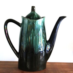 May include: A ceramic coffee pot with a dark green and black glaze. The pot features a curved handle, a long spout, and a lid with a small knob. The glaze has a glossy finish. The pot is on a wooden surface.