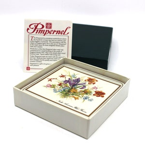 Könnte beinhalten: Ein Set Pimpernel-Untersetzer in einer weißen Box. Jeder Untersetzer zeigt ein Blumendesign mit lila Iris, orangefarbenen und blauen Blumen. Die Box enthält auch eine Karte mit dem Pimpernel-Logo und Text. Eine dunkelgrüne Box ist ebenfalls sichtbar.