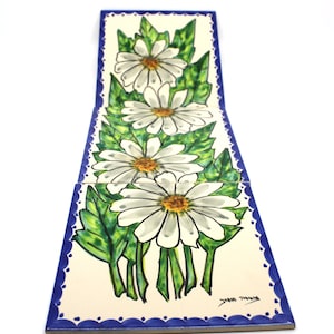 May include: Four white daisy flowers with yellow centres are painted on a set of four ceramic tiles. The tiles are edged in blue and have a scalloped border. The tiles are signed "Juber Tamayo".