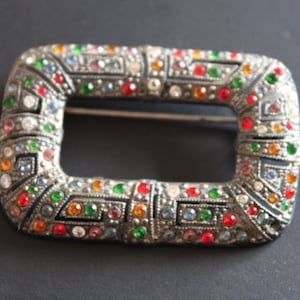 Peut inclure: Boucle rectangulaire argentée ornée de pierres précieuses colorées. La boucle est ornée de pierres rouges, vertes, bleues, oranges et transparentes. Elle présente une ouverture centrale et une épingle au dos, pour ceintures ou accessoires.