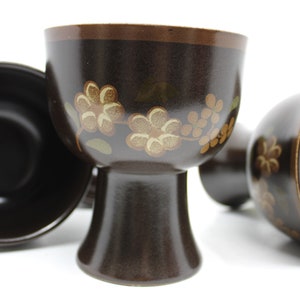 May include: Set of four brown ceramic cups with a floral design. The cups have a wide base and a narrow top.