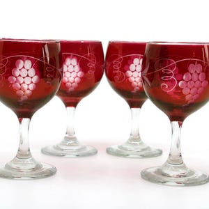 May include: Four ruby red wine glasses with clear stems. Each glass has an etched design of a grape cluster and vines. The glasses are arranged on a white surface, ideal for serving wine.