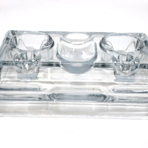 May include: A clear glass inkwell with three wells for holding ink. The inkwell has a rectangular base and is made of thick glass.