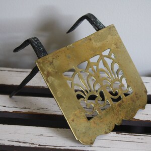May include: A brass and iron fireplace grate with a decorative cutout design. The grate has two iron legs that extend out from the sides.