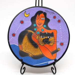 Peut inclure: Une assiette ronde en céramique avec un fond violet et une femme aux longs cheveux noirs tenant un chat. La femme porte un collier turquoise et le chat a une fourrure noire et orange. L'assiette a une lune croissante et des étoiles en arrière-plan. L'assiette est signée "ZUTCHIKA INC."