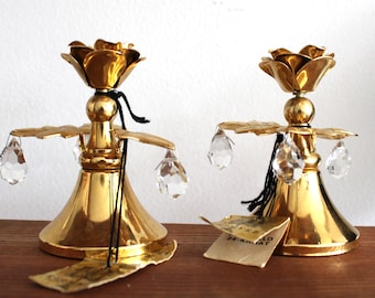May include: Two gold-colored candle holders with a floral design at the top. Each candle holder has a round base, a central stem, and crystal-like pendants hanging from the sides. A tag is attached to one of the candle holders.