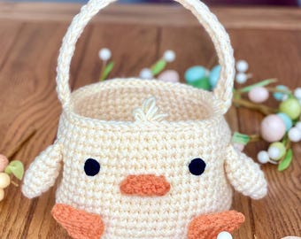 May include: A crocheted chick basket with a cream-colored body, orange beak and feet, and black eyes. The basket has a handle and is perfect for holding small items.
