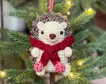 May include: A crocheted hedgehog ornament with a red scarf and white pom-poms on its feet. The ornament is hanging from a red and white striped string on a small artificial Christmas tree.