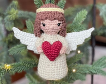 May include: A crocheted angel ornament with white wings, a brown head, and a pink face. The angel is holding a red heart in its hands.