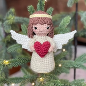 May include: A crocheted angel ornament with white wings, a brown head, and a pink face. The angel is holding a red heart in its hands.