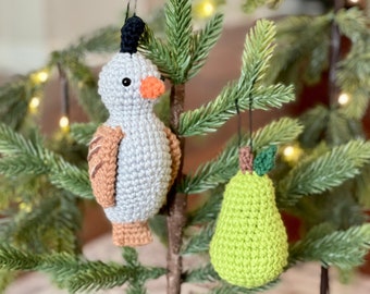 May include: Two crocheted ornaments hanging on a small artificial Christmas tree. One ornament is a gray bird with an orange beak and black eyes. The other ornament is a green pear with a brown stem and green leaves.