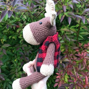 May include: A brown crocheted moose wearing a red and black plaid vest. The moose has large antlers and a black button eye.