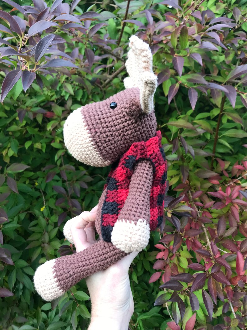 May include: A brown crocheted moose wearing a red and black plaid vest. The moose has large antlers and a black button eye.