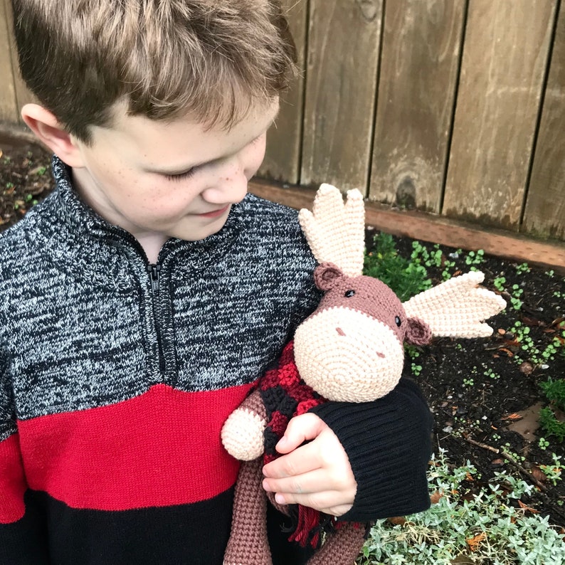 May include: A child holds a crocheted moose toy wearing a red and black plaid scarf. The moose is brown with a cream-colored face and large, floppy ears.