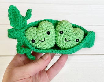 May include: A handmade crochet pea pod with two smiling peas. The peas are heart-shaped and light green, with black eyes and a stitched smile. The pod is also green, with a textured, leafy design.