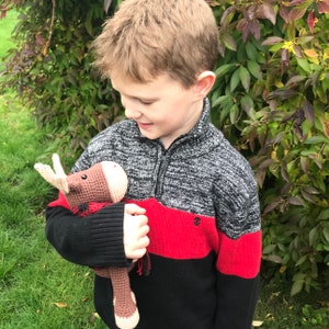 May include: A young person wearing a black and red sweater holds a brown and tan crocheted moose toy. The toy has a red and black plaid scarf.