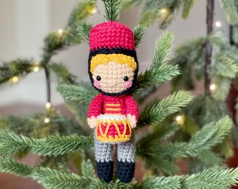 May include: A crocheted toy soldier ornament with a red hat, a red and white drum, and a yellow face. The ornament is hanging on a green Christmas tree.
