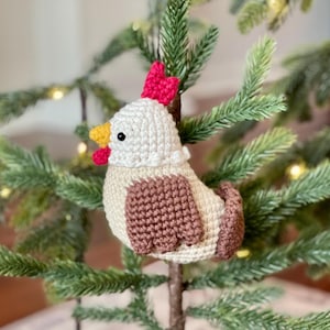 May include: A crocheted chicken ornament with a white body, brown wings, and a red comb. The ornament is hanging on a green Christmas tree.