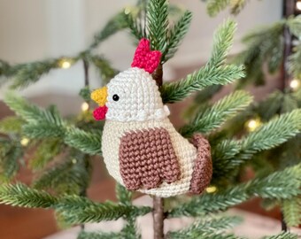 May include: A crocheted chicken ornament with a white body, brown wings, and a red comb. The ornament is hanging on a green Christmas tree.