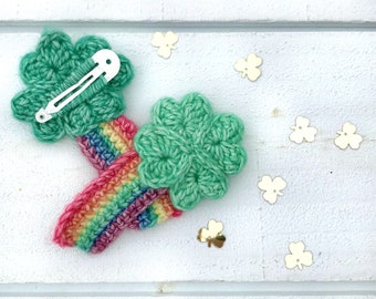 May include: Two green crocheted four-leaf clover hair clips with rainbow stripes. The clips are on a white wooden surface with gold glitter shamrocks.