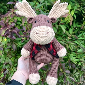 May include: A brown crocheted moose with a red and black plaid scarf. The moose is sitting on a hand and has a white face.