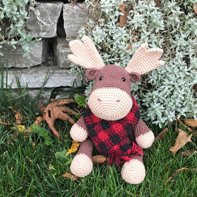 May include: A brown crocheted moose wearing a red and black plaid scarf. The moose is sitting on a bed of green grass and brown leaves.