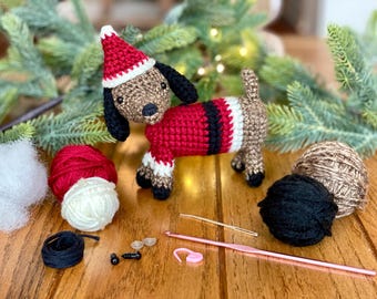 May include: A crocheted dachshund wearing a red and white Santa hat and a red and black sweater. The dachshund is surrounded by yarn balls, a crochet hook, and other supplies for making the toy.