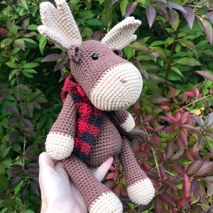 May include: A brown crocheted moose with a red and black plaid scarf. The moose is sitting on a person's hand.