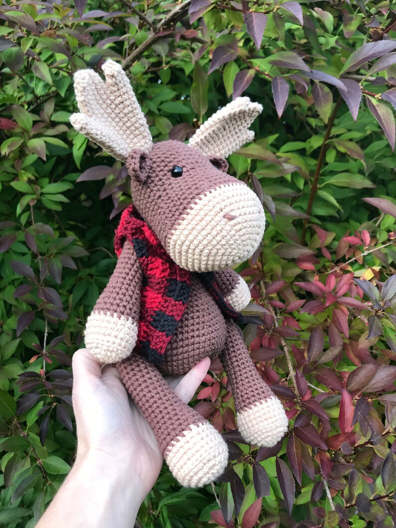 May include: A brown crocheted moose with a red and black plaid scarf. The moose is sitting on a person's hand.