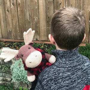 May include: A crocheted moose toy wearing a red and black plaid scarf. The moose is brown with a cream-colored face and is being held by a child.