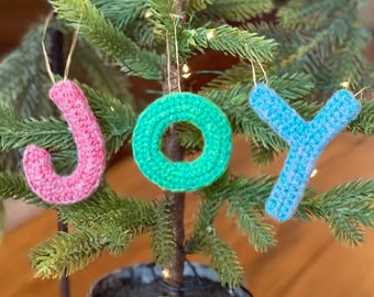 May include: Crocheted ornaments spelling out the word "JOY" in pink, green, and blue. The ornaments are hanging on a small artificial Christmas tree.