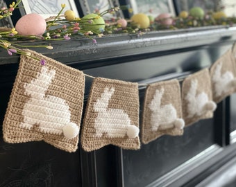 May include: A brown crocheted bunny garland with white bunny silhouettes. The garland is hanging on a black mantelpiece.