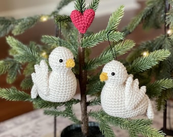 May include: Two white crocheted doves with yellow beaks and black eyes hanging from a small artificial Christmas tree with a red crocheted heart ornament.