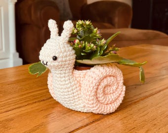 May include: A handmade crochet snail planter in cream and light pink. The snail has a spiral shell and holds a small potted plant with green leaves and small red flowers. The snail is sitting on a wooden surface.