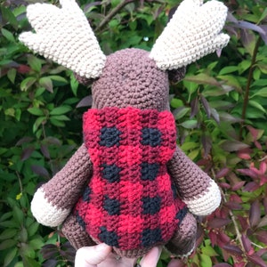 May include: A brown crocheted moose wearing a red and black plaid hooded sweater. The moose is standing on a hand.
