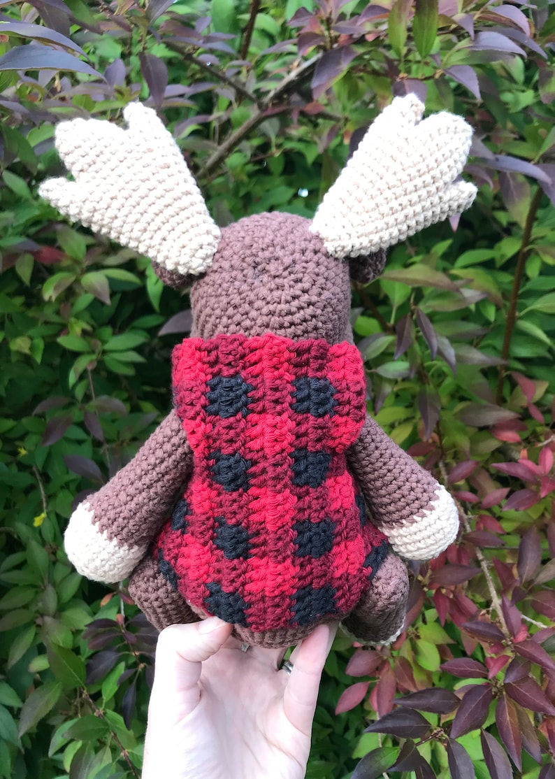May include: A brown crocheted moose wearing a red and black plaid hooded sweater. The moose is standing on a hand.