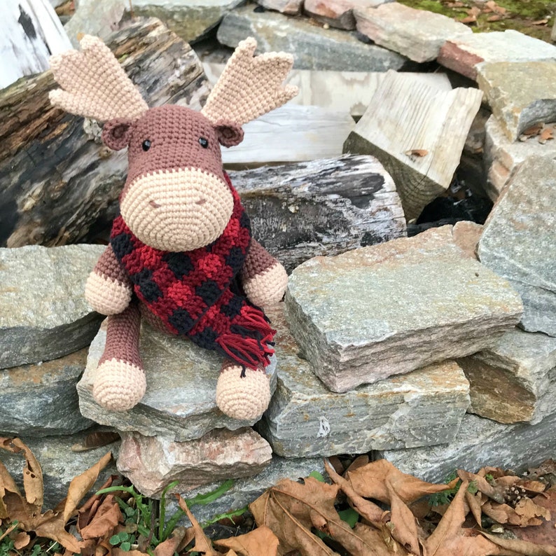May include: A brown crocheted moose wearing a red and black plaid scarf, sitting on a pile of rocks.
