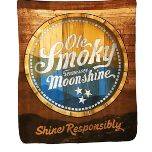 Pu&ograve; includere: Uno sfondo in legno marrone con un logo giallo, blu e bianco per Ole Smoky Tennessee Moonshine. Il logo &egrave; in un cerchio con una texture a grana di legno e il testo "Shine Responsibly" &egrave; sotto il logo.