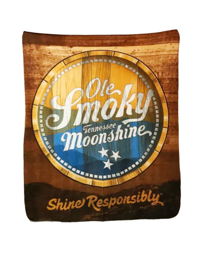 Pu&ograve; includere: Uno sfondo in legno marrone con un logo giallo, blu e bianco per Ole Smoky Tennessee Moonshine. Il logo &egrave; in un cerchio con una texture a grana di legno e il testo "Shine Responsibly" &egrave; sotto il logo.